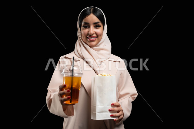 النظر الى الكاميرا بايماءات الفرح والسعادة، قضاء أوقات ممتعة أثناء العطلة، الاستعداد للذهاب الى السينما لرؤية الفيلم المفضل، بورتريه مقرب لامرأة عربية خليجية سعودية محجبة ترتدي عباءه بيج تحمل كيس فوشار وكأس من المشروب الغازي، خلفية سوداء