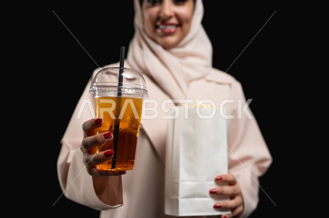 الذهاب الى السينما لرؤية الفيلم المفضل، ايماءات الفرح والسعادة، قضاء أوقات ممتعة أثناء العطلة، بورتريه مقرب ليدين امرأة عربية خليجية سعودية محجبة ترتدي عباءه بيج تحمل كيس فوشار وكأس من المشروب الغازي، خلفية سوداء