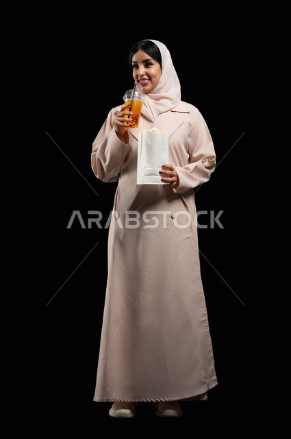 التسلية والترفيه بقضاء أوقات ممتعة، الاستمتاع بمشاهدة الأفلام داخل السينما، بورتريه لامرأة عربية خليجية سعودية محجبة ترتدي عباءه بيج وتحمل كيس فوشار وكأس من المشروب الغازي، ايماءات السعادة والسرور لمتابعة الفيلم المفضل، الجسم بالطول الكامل، خلفية سوداء