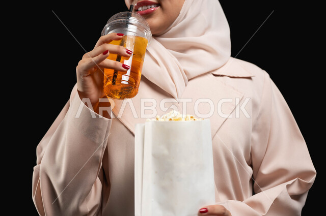 الترفيه والتسلية بقضاء أوقات ممتعة، الاستمتاع بمشاهدة الأفلام داخل السينما، بورتريه مقرب لامرأة عربية خليجية سعودية محجبة ترتدي عباءه بيج وتحمل كيس فوشار وكأس من المشروب الغازي، متابعة الفيلم المفضل، خلفية سوداء