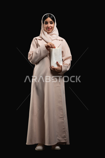 Enjoying the weekend, spending enjoyable times watching movies, cinema, and series, a portrait of a smiling, naked Saudi Gulf woman wearing a hijab and abaya, holding a bag of popcorn, standing straight and looking at the camera with expressions of pleasure and happiness, a full-body portrait, black background.
