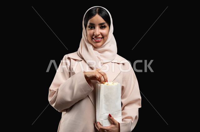 تناول الفوشار والتسالي اثناء متابعة الأفلام المسلية، بورتريه مقرب لإمرأة عربية خليجية سعودية مبتسمة ترتدي الحجاب والعباءة تقوم بالأنشطة الترفيهيه في عطلة نهاية الإسبوع، قضاء وقت ممتع في السينما، النظر الى الكاميرا بتعابير السرور والسعادة، خلفية سوداء