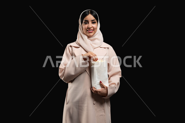 تناول الفوشار والتسالي اثناء متابعة الأفلام المسلية، بورتريه مقرب لإمرأة عربية خليجية سعودية مبتسمة ترتدي الحجاب والعباءة تقوم بالأنشطة الترفيهيه في عطلة نهاية الإسبوع، قضاء وقت ممتع في السينما، النظر الى الكاميرا بتعابير السرور والسعادة، خلفية سوداء