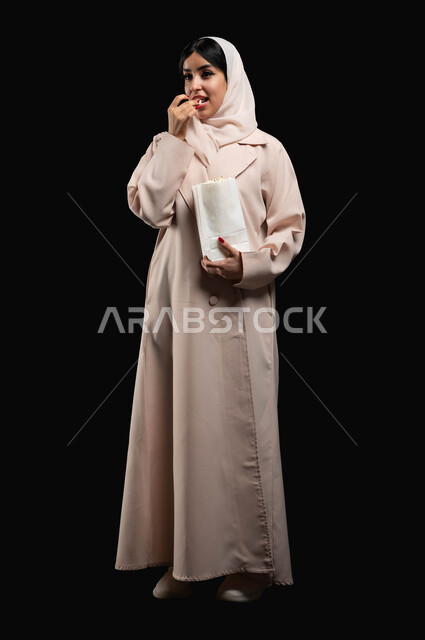 الوقوف باستقامة والنظر الى شيء ما بتعابير السرور والاندماج، قضاء وقت ممتع بمتابعة الأفلام والسينما والمسلسلات، الاستمتاع في عطلة نهاية الإسبوع، بورتريه لشابة عريية خليجية سعودية مبتسمة ترتدي الحجاب والعباءة تقوم بتناول الفوشار، صورة لكامل الجسم، خلفية سوداء