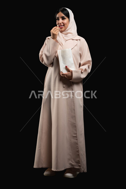 الوقوف باستقامة والنظر الى شيء ما بتعابير السرور والاندماج، قضاء وقت ممتع بمتابعة الأفلام والسينما والمسلسلات، الاستمتاع في عطلة نهاية الإسبوع، بورتريه لشابة عريية خليجية سعودية مبتسمة ترتدي الحجاب والعباءة تقوم بتناول الفوشار، صورة لكامل الجسم، خلفية سوداء