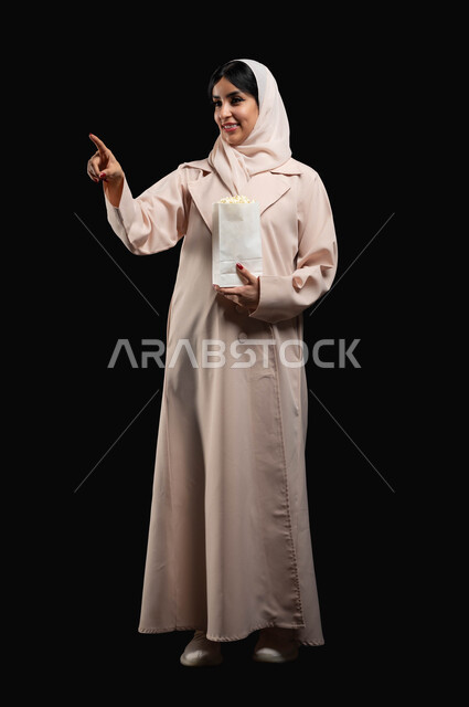 الاشارة الى شيء ما بتعابير التركيز والاندماج، قضاء وقت ممتع بمتابعة الأفلام والسينما والمسلسلات، الاستمتاع في عطلة نهاية الإسبوع، بورتريه لشابة عريية خليجية سعودية مبتسمة ترتدي الحجاب والعباءة تحمل كيس الفوشار تقف باستقامة، صورة لكامل الجسم، خلفية سوداء