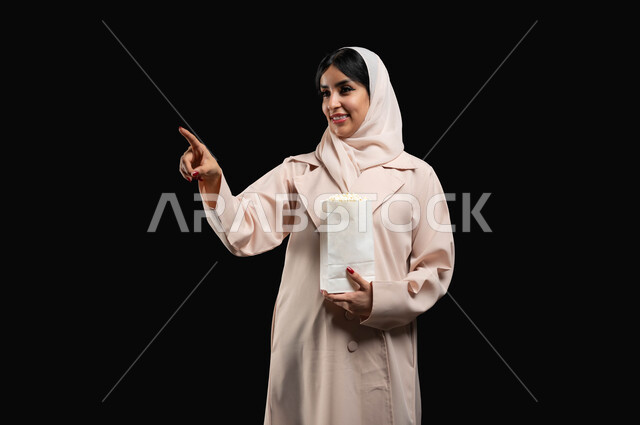 الاشارة الى شيء ما بتعابير السرور والاندماج، قضاء وقت ممتع بمتابعة الأفلام والسينما والمسلسلات، الاستمتاع في عطلة نهاية الإسبوع، بورتريه مقرب لإمرأة عربية خليجية سعودية مبتسمة ترتدي الحجاب والعباءة تحمل كيس الفوشار تقف باستقامة، خلفية سوداء