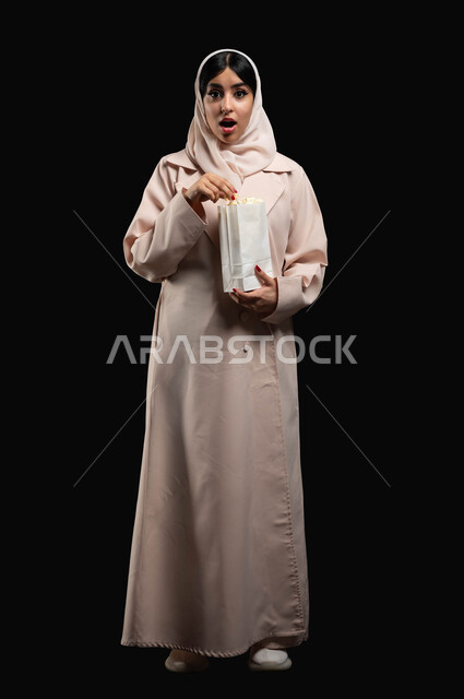 النظر الى الكاميرا بتعابير الاندهاش والصدمة والذهول، قضاء وقت ممتع بمتابعة أفلام الرعب والأكشن، الاستمتاع في عطلة نهاية الإسبوع، بورتريه لشابة عريية خليجية سعودية ترتدي الحجاب والعباءة تحمل كيس الفوشار تقف باستقامة، صورة الجسم بالطول الكامل، خلفية سوداء