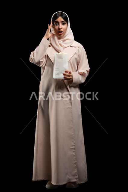 Eating popcorn and snacks while watching suspense and thriller movies, portrait of a Saudi Gulf Arab young woman wearing the hijab and abaya, placing her hand on her head with an expression of astonishment and shock, standing up straight and looking at the camera, spending a good time at the cinema, full body portrait, black background