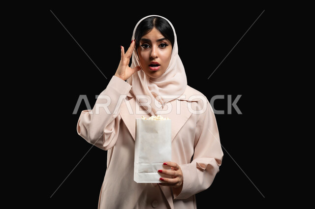 تناول الفوشار والتسالي اثناء متابعة أفلام التشويق والاثارة، بورتريه مقرب لإمرأة عربية خليجية سعودية ترتدي الحجاب والعباءة تضع يدها على رأسها بتعابير الاندهاش والصدمة والذهول، الوقوف باستقامة والنظر الى الكاميرا، قضاء وقت ممتع في السينما، خلفية سوداء