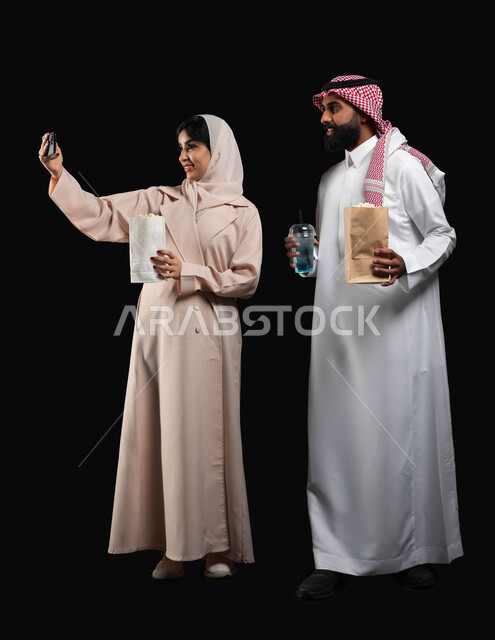 قضاء اوقات ممتعة وتناول الفوشار والمشروبات اثناء متابعة الافلام والمسلسلات، توثيق اللحظات العائلية السعيدة، بورتريه لزوجين عربيين خليجيين سعوديين يلتقطان صور سيلفي باستخدام الهاتف المحمول، الاستعداد لمشاهدة فيلم في السينما، صورة الجسم بالكامل، خلفية سوداء