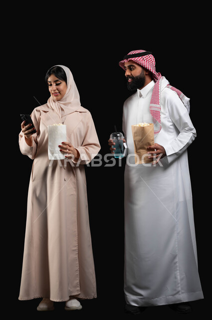 استخدام الأجهزة التقنية الحديثة، بورتريه لإمرأة عربية خليجية سعودية مبتسمة ترتدي الحجاب والعباءة تمسك الهاتف المحمول تقف بجانب زوجها، إحضار الفوشار والمشروبات والاستعداد لمشاهدة فيلم في السينما، قضاء أوقات ممتعة في العطلة، صورة الجسم بالكامل، خلفية سوداء