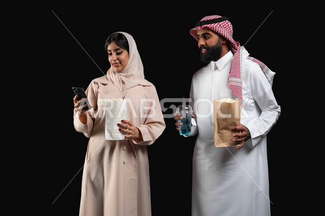 إحضار الفوشار والمشروبات الباردة والاستعداد لمشاهدة فيلم في السينما، قضاء أوقات ممتعة في عطلة نهاية الاسبوع، استخدام الأجهزة التقنية الحديثة، بورتريه مقرب لإمرأة عربية خليجية سعودية مبتسمة ترتدي الحجاب والعباءة تمسك الهاتف المحمول تقف بجانب زوجها، خلفية سوداء