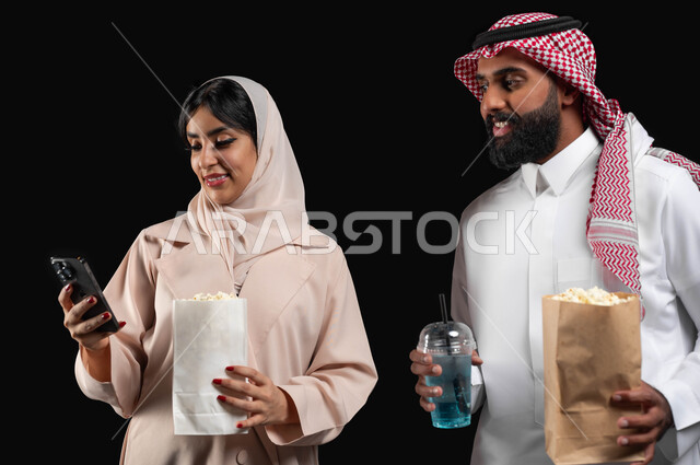 إحضار الفوشار والمشروبات الباردة والاستعداد لمشاهدة فيلم في السينما، قضاء أوقات ممتعة في عطلة نهاية الاسبوع، استخدام الأجهزة التقنية الحديثة، بورتريه مقرب لإمرأة عربية خليجية سعودية مبتسمة ترتدي الحجاب والعباءة تمسك الهاتف المحمول تقف بجانب زوجها، خلفية سوداء