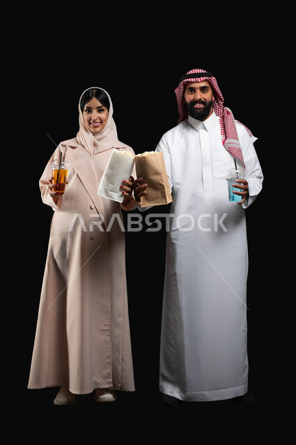 قضاء وقت ممتع في السينما، تناول الفوشار والمشروبات الباردة اثناء متابعة الأفلام والمسلسلات، بورتريه لزوجيين عربيين خليجيين سعوديين مبتسميين ينظران الى الكاميرا بتعابير السرور والسعادة، الاستمتاع بالأنشطة العائلية الترفيهيه، صورة لكامل الجسم، خلفية سوداء