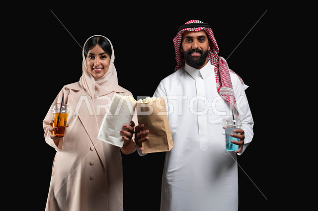 Enjoying family entertainment activities on the weekend, spending a good time at the movies, eating popcorn and cold drinks while watching movies and series, a close-up portrait of a smiling Gulf Arab-Saudi couple looking at the camera with expressions of pleasure and happiness, black background.