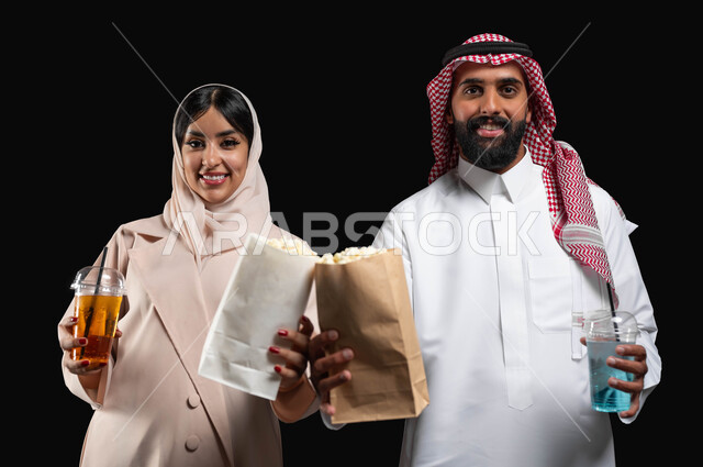 فعاليات وأنشطة عائلية ترفيهيه، قضاء وقت ممتع في السينما، تناول الفوشار والمشروبات الباردة اثناء متابعة الأفلام والمسلسلات في عطلة نهاية الاسبوع، بورتريه مقرب لزوجيين عربيين خليجيين سعوديين مبتسميين ينظران الى الكاميرا بتعابير السرور والسعادة، خلفية سوداء