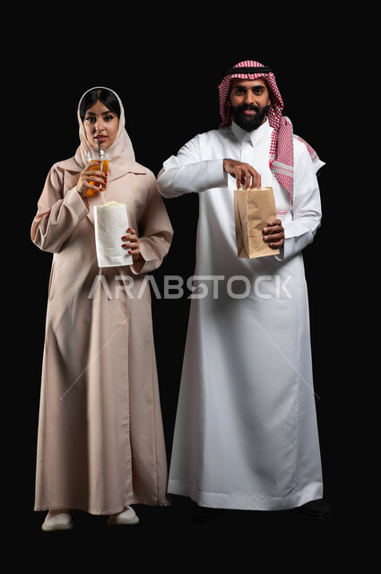Spending a good time at the cinema, eating popcorn and cold drinks while watching movies and series, a portrait of a smiling Gulf Arab-Saudi couple looking at the camera with expressions of pleasure and happiness, enjoying recreational family activities, full body portrait, black background