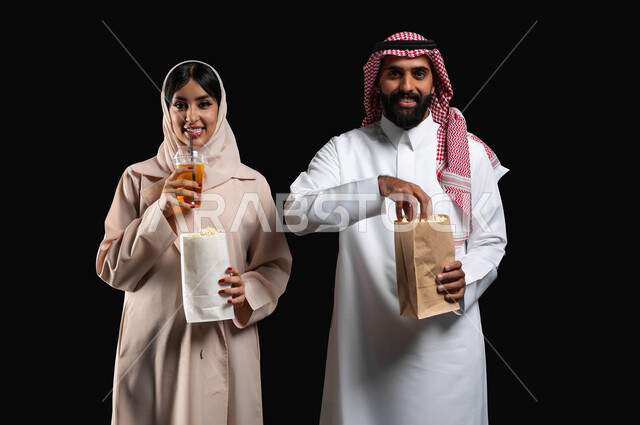 Enjoying family entertainment activities on the weekend, spending a good time at the movies, eating popcorn and cold drinks while watching movies and series, a close-up portrait of a smiling Gulf Arab-Saudi couple looking at the camera with expressions of pleasure and happiness, black background.