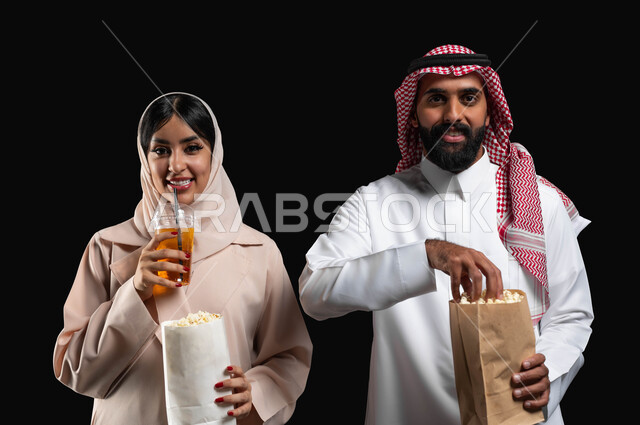 Family entertainment events and activities, spending a good time at the movies, eating popcorn and cold drinks while watching movies and series on the weekend, a close-up portrait of a smiling Gulf Arab-Saudi couple looking at the camera with expressions of pleasure and happiness, a black background.