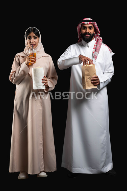 Spending a good time at the cinema, eating popcorn and cold drinks while watching movies and series, a portrait of a smiling Gulf Arab-Saudi couple looking at the camera with expressions of pleasure and happiness, enjoying recreational family activities, full body portrait, black background
