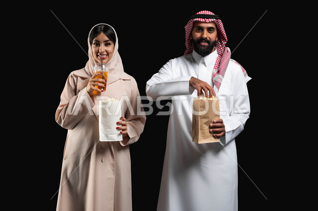 Enjoying family entertainment activities on the weekend, spending a good time at the movies, eating popcorn and cold drinks while watching movies and series, a close-up portrait of a smiling Gulf Arab-Saudi couple looking at the camera with expressions of pleasure and happiness, black background.