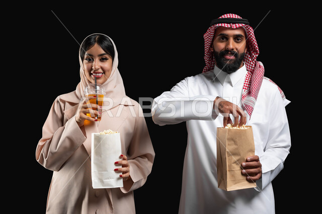 فعاليات وأنشطة عائلية ترفيهيه، قضاء وقت ممتع في السينما، تناول الفوشار والمشروبات الباردة اثناء متابعة الأفلام والمسلسلات في عطلة نهاية الاسبوع، بورتريه مقرب لزوجيين عربيين خليجيين سعوديين مبتسميين ينظران الى الكاميرا بتعابير السرور والسعادة، خلفية سوداء
