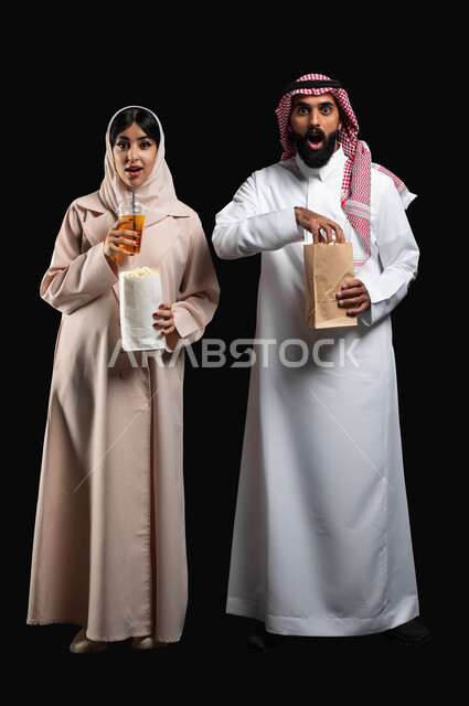 قضاء وقت ممتع في السينما، تناول الفوشار والمشروبات الباردة اثناء متابعة أفلام التشويق والاثارة، بورتريه لزوجيين عربيين خليجيين سعوديين ينظران الى الكاميرا بتعابير الاندهاش والصدمة، الاستمتاع بالأنشطة العائلية الترفيهيه في العطلة، صورة لكامل الجسم، خلفية سوداء