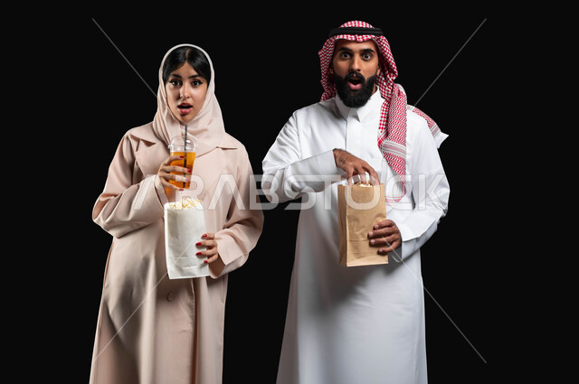 Eating popcorn and cold drinks while watching suspense and excitement films, a close-up portrait of a Saudi Gulf Arab couple looking at the camera with expressions of amazement, shock and astonishment, spending a good time at the cinema, enjoying recreational family activities on vacation, a black background