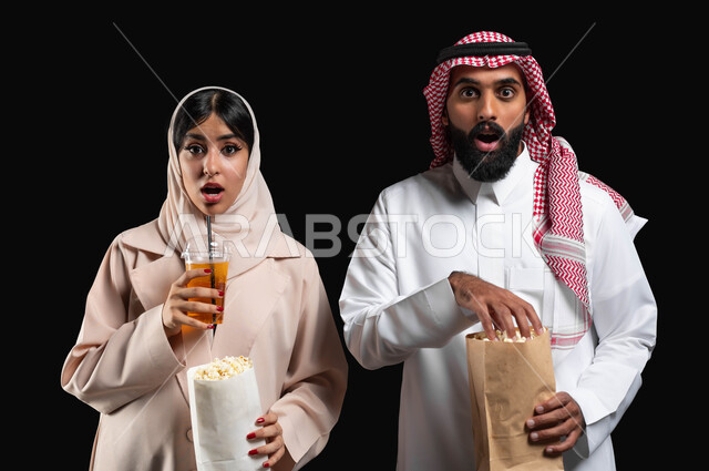 تناول الفوشار والمشروبات الباردة اثناء متابعة أفلام التشويق والاثارة، بورتريه مقرب لزوجيين عربيين خليجيين سعوديين ينظران الى الكاميرا بتعابير الاندهاش والصدمة والذهول، قضاء وقت ممتع في السينما، الاستمتاع بالأنشطة العائلية الترفيهيه في العطلة، خلفية سوداء
