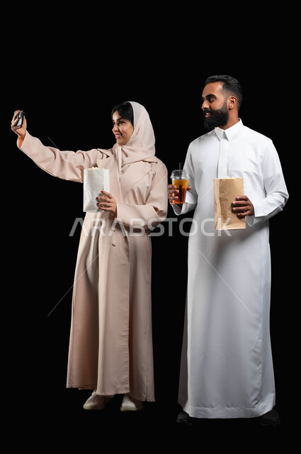 قضاء اوقات ممتعة وتناول الفوشار والمشروبات اثناء متابعة الافلام والمسلسلات، توثيق اللحظات العائلية السعيدة، بورتريه لزوجين عربيين خليجيين سعوديين يلتقطان صور سيلفي باستخدام الهاتف المحمول، الاستعداد لمشاهدة فيلم في السينما، صورة الجسم بالكامل، خلفية سوداء