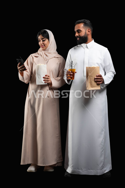 استخدام الأجهزة التقنية الحديثة، بورتريه لإمرأة عربية خليجية سعودية مبتسمة ترتدي الحجاب والعباءة تمسك الهاتف المحمول تقف بجانب زوجها،  إحضار الفوشار والمشروبات والاستعداد لمشاهدة فيلم في السينما، قضاء أوقات ممتعة في العطلة، صورة الجسم بالكامل، خلفية سوداء