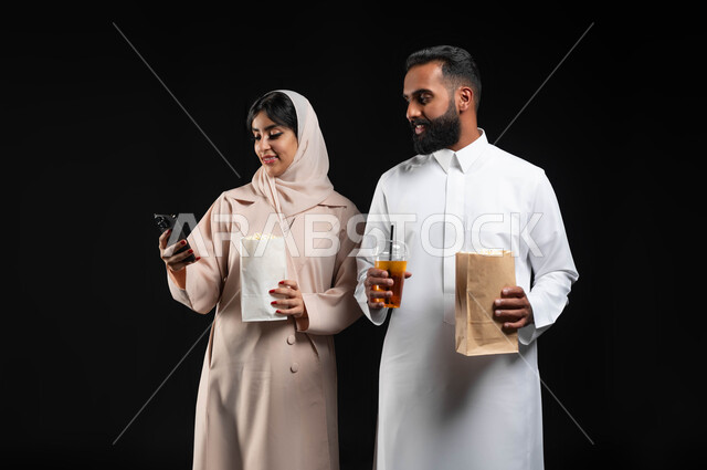 استخدام الأجهزة التقنية الحديثة، بورتريه مقرب لإمرأة عربية خليجية سعودية مبتسمة ترتدي الحجاب والعباءة تمسك الهاتف المحمول تقف بجانب زوجها،  إحضار الفوشار والمشروبات والاستعداد لمشاهدة فيلم في السينما، قضاء أوقات ممتعة في عطلة نهاية الاسبوع، خلفية سوداء