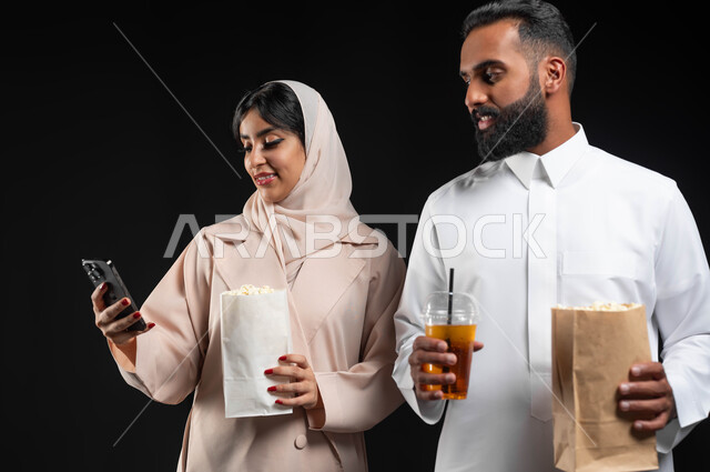 استخدام الأجهزة التقنية الحديثة، بورتريه مقرب لإمرأة عربية خليجية سعودية مبتسمة ترتدي الحجاب والعباءة تمسك الهاتف المحمول تقف بجانب زوجها،  إحضار الفوشار والمشروبات والاستعداد لمشاهدة فيلم في السينما، قضاء أوقات ممتعة في عطلة نهاية الاسبوع، خلفية سوداء