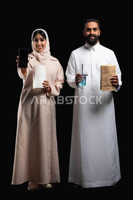 عرض شاشة هاتف محمول سوداء فارغة، بورتريه لإمرأة عربية خليجية سعودية مبتسمة ترتدي الحجاب والعباءة تمسك الجوال تقف بجانب زوجها، إحضار الفوشار والمشروبات والاستعداد لمشاهدة فيلم في السينما، قضاء أوقات ممتعة في العطلة، صورة الجسم بالطول الكامل، خلفية سوداء