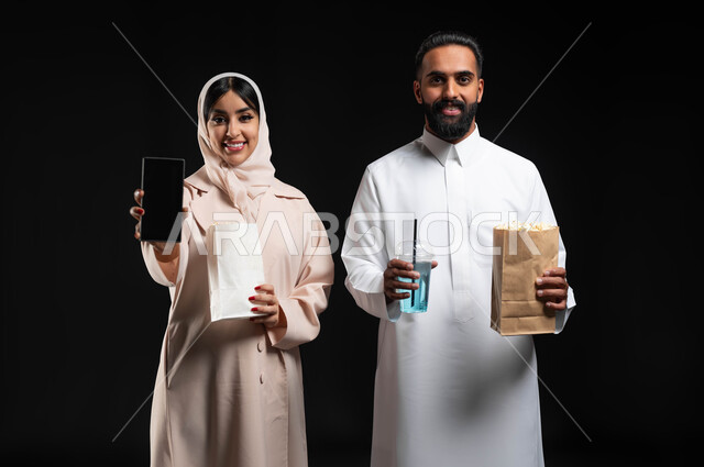 قضاء أوقات ممتعة في العطلة، استخدام الاجهزة التقنية، عرض شاشة هاتف محمول سوداء فارغة، بورتريه مقرب لإمرأة عربية خليجية سعودية مبتسمة ترتدي الحجاب والعباءة تمسك الجوال تقف بجانب زوجها، إحضار الفوشار والمشروبات والاستعداد لمشاهدة فيلم في السينما، خلفية سوداء