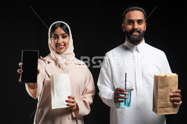 استخدام الاجهزة التقنية، عرض شاشة هاتف محمول سوداء فارغة، بورتريه مقرب لإمرأة عربية خليجية سعودية مبتسمة ترتدي الحجاب والعباءة تمسك الجوال تقف بجانب زوجها، إحضار الفوشار والمشروبات والاستعداد لمشاهدة فيلم في السينما، قضاء أوقات ممتعة في العطلة، خلفية سوداء