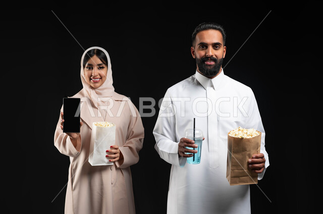 Spending fun times on vacation, using technical devices, showing a blank black mobile phone screen, a close-up portrait of a smiling Saudi Gulf Arab woman wearing the hijab and abaya, holding the mobile phone, standing next to her husband, bringing popcorn and drinks and getting ready to watch a movie in the cinema, black background