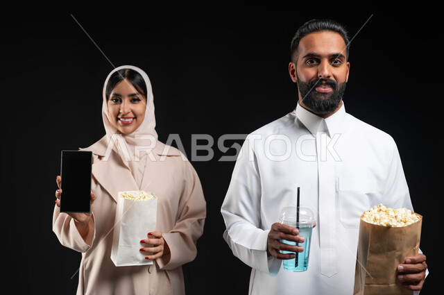 استخدام الاجهزة التقنية، عرض شاشة هاتف محمول سوداء فارغة، بورتريه مقرب لإمرأة عربية خليجية سعودية مبتسمة ترتدي الحجاب والعباءة تمسك الجوال تقف بجانب زوجها، إحضار الفوشار والمشروبات والاستعداد لمشاهدة فيلم في السينما، قضاء أوقات ممتعة في العطلة، خلفية سوداء