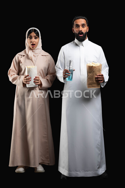 مشاهدة أفلام الرعب والأكشن، قضاء أوقات ممتعة والاستمتاع في عطلة نهاية الإسبوع، بورتريه لزوجيين عربيين خليجيين سعوديين ينظران الى الكاميرا بتعابير الاندهاش والصدمة والذهول، تناول الفوشار والمشروبات الباردة في السينما، صورة الجسم بالطول الكامل، خلفية سوداء