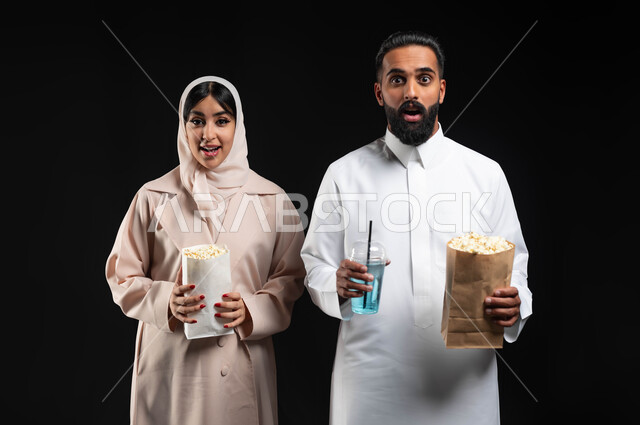 النظر الى الكاميرا بتعابير الاندهاش والصدمة والذهول، قضاء أوقات ممتعة بمتابعة أفلام الرعب والأكشن، الاستمتاع في عطلة نهاية الإسبوع، بورتريه مقرب لزوجيين عربيين خليجيين سعوديين يقومان بتناول الفوشار والمشروبات الباردة، أجواء اسرية سعيدة، خلفية سوداء