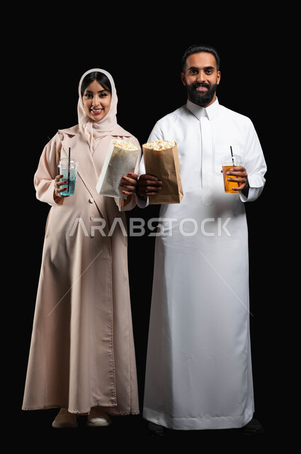 تناول الفوشار والمشروبات الباردة اثناء متابعة الأفلام والمسلسلات، بورتريه لزوجيين عربيين خليجيين سعوديين مبتسميين ينظران الى الكاميرا بتعابير السرور، الاستمتاع بالأنشطة العائلية الترفيهيه في العطلة، قضاء وقت ممتع في السينما، صورة لكامل الجسم، خلفية سوداء