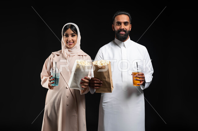 الاستمتاع بالأنشطة العائلية الترفيهيه في عطلة نهاية الاسبوع، قضاء وقت ممتع في السينما، تناول الفوشار والمشروبات الباردة اثناء متابعة الأفلام والمسلسلات، بورتريه مقرب لزوجيين عربيين خليجيين سعوديين مبتسميين ينظران الى الكاميرا بتعابير السرور والسعادة، خلفية سوداء