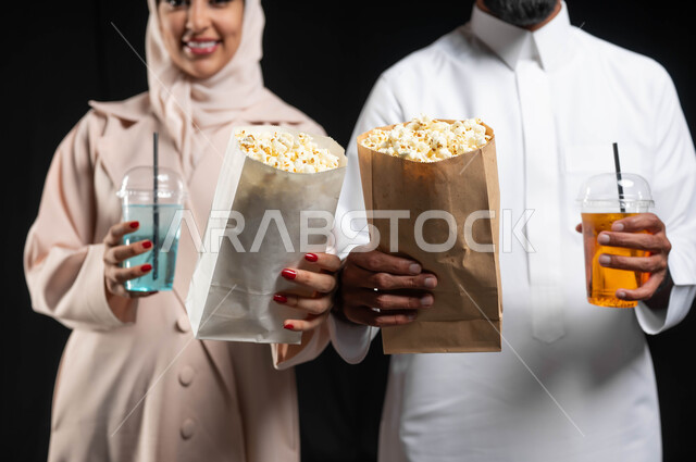 الاستمتاع بالأنشطة العائلية الترفيهيه في عطلة نهاية الاسبوع، قضاء وقت ممتع في السينما، تناول الفوشار والمشروبات الباردة اثناء متابعة الأفلام والمسلسلات، بورتريه مقرب لزوجيين عربيين خليجيين سعوديين مبتسميين ينظران الى الكاميرا بتعابير السرور والسعادة، خلفية سوداء