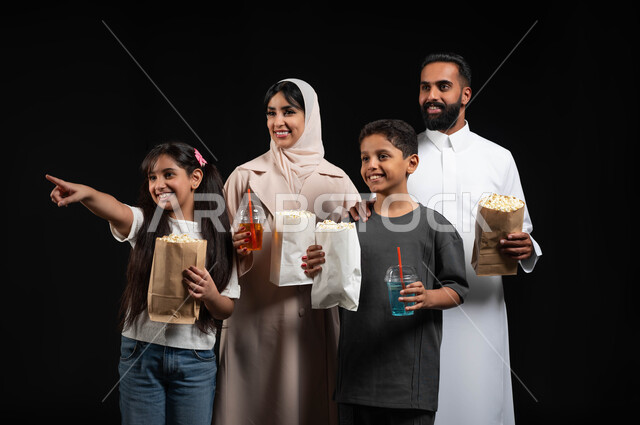 الاشارة والنظر الى شيء ما بتعابير السرور، بورتريه مقرب لعائلة عربية خليجية سعودية تقضي أوقات ممتعة في عطلة نهاية الاسبوع، أنشطة وفعاليات عائلية مسلية، إحضار الفوشار والمشروبات الباردة، الاجواء الاسرية السعيدة، الاستعداد لمشاهدة فيلم في السينما، خلفية سوداء