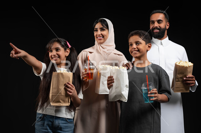 Pointing and looking at something with an expression of pleasure, a close-up portrait of a Saudi Gulf Arab family spending enjoyable times on the weekend, entertaining family activities and events, bringing popcorn and cold drinks, happy family atmosphere, preparing to watch a movie in the cinema, black background