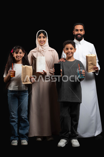 عرض شاشة هاتف محمول سوداء فارغة، أنشطة وفعاليات عائلية مسلية، إحضار الفوشار والتسالي والمشروبات الباردة، استخدام الاجهزة التقنية الحديثة، بورتريه لعائلة عربية خليجية سعودية تستعد لمشاهدة فيلم في السينما بتعابير السرور، صورة الجسم بالطول الكامل، خلفية سوداء