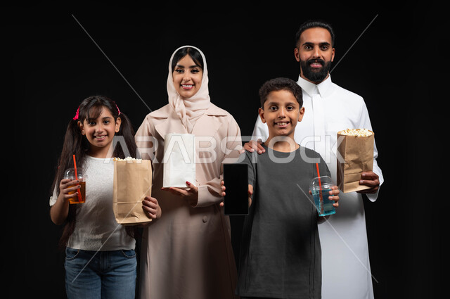 Displaying the mobile screen, gestures of happiness and pleasure, using modern technology, a happy family atmosphere, a portrait of a Saudi Gulf Arab family, watching movies and series, recreational weekend activities, enjoying popcorn and cold drinks, black background