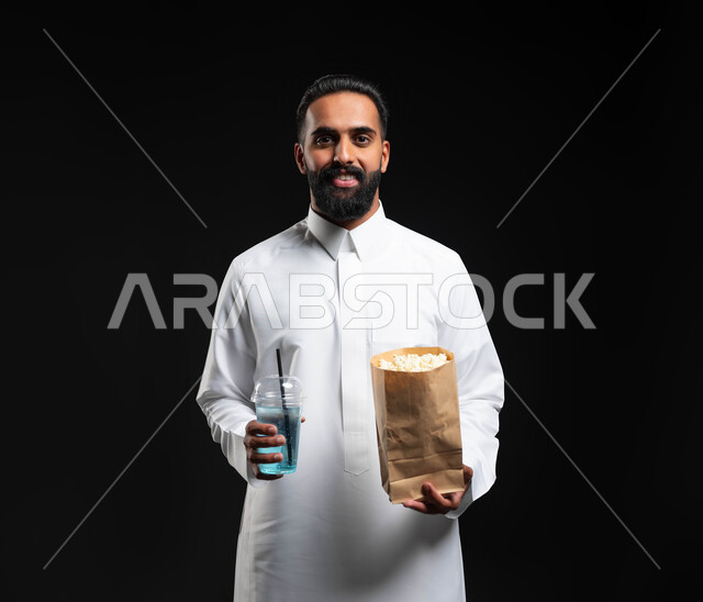 متابعة برامج التلفاز، النظر إلى الكاميرا بايماءات السعادة والسرور، تناول الفوشار والمشروبات الباردة، انشطة ترفيهية منزلية، بورتريه لرجل عربي خليجي سعودي يرتدي الثوب التقليدي الأبيض، الاستمتاع بمشاهدة الأفلام والمسلسلات، قضاء وقت ممتع في المنزل، خلفية سوداء