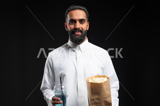 النظر إلى الكاميرا بايماءات السعادة والسرور، تناول الفوشار والمشروبات الباردة، انشطة ترفيهية منزلية، بورتريه مقرب لرجل عربي خليجي سعودي يرتدي الثوب التقليدي الأبيض، الاستمتاع بمشاهدة الأفلام والمسلسلات، متابعة برامج التلفاز، قضاء وقت ممتع في المنزل، خلفية سوداء